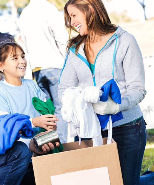 Mother and daughter smile as they place canned food and clothing in donation box at outdoor community food and clothing drive. Volunteers are helping people out in the background.